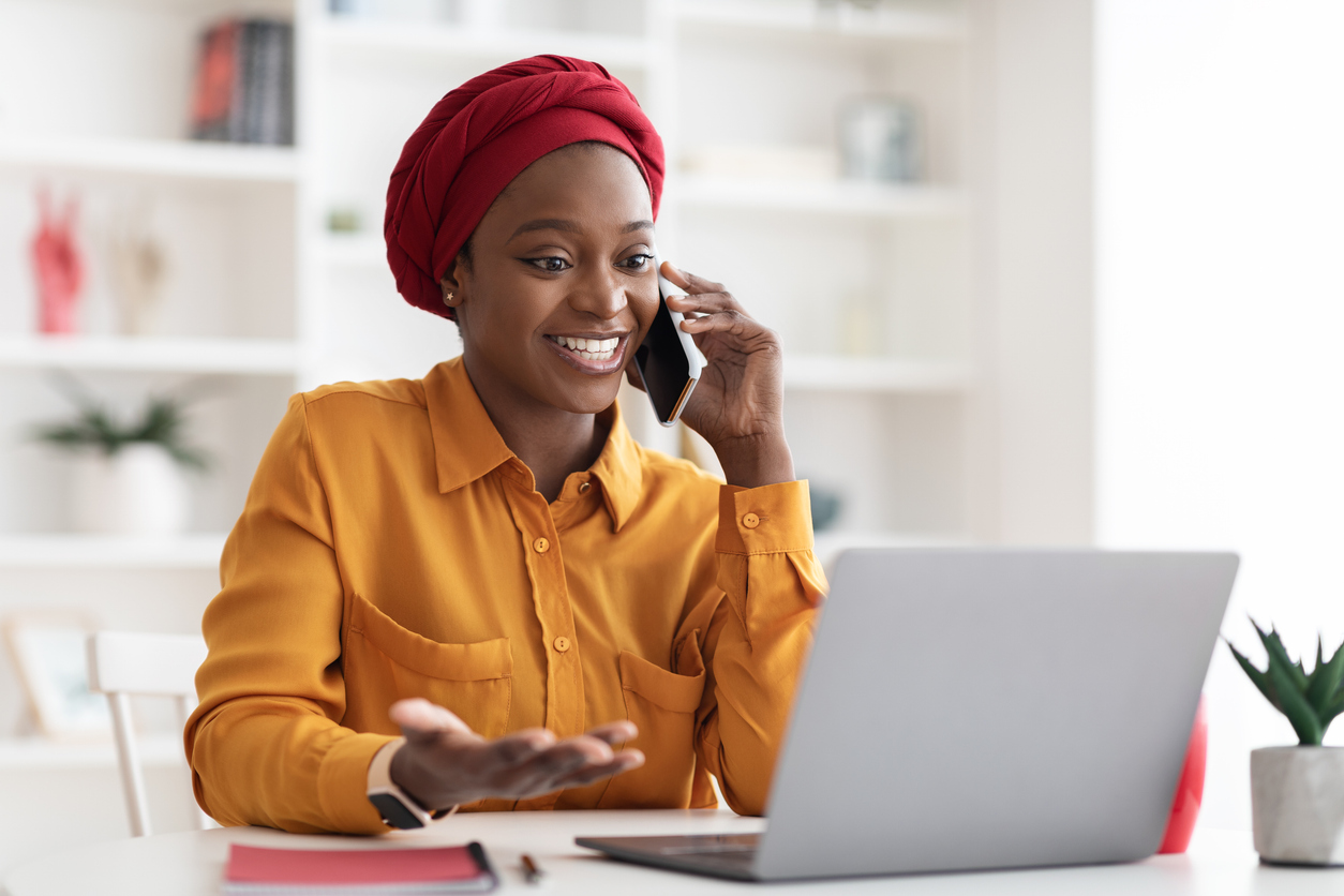 Positive young muslim black lady with red turban on her head and casual outfit working on laptop at office, looking at computer screen and talking on cell phone with business partner, copy space